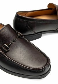 Brązowe skórzane mokasyny o gładkiej fakturze, kontrastowym przeszyciu i dekoracyjnym metalowym łańcuchu na wierzchu. Niska gumowa podeszwa.