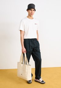 Camiseta blanca de manga corta con logo, pantalones negros con cintura elástica, bolso tote acolchado color crema y zapatos negros con detalles en rosa sobre un suelo amarillo.