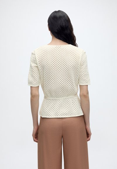 Γυναίκα με μακριά σκούρα μαλλιά που φοράει μαλακό κρεμ κοντομάνικο τοπ πλεχτό και καφέ παντελόνα ψηλόμεση με φαρδιά πόδια, στέκεται με την πλάτη στην κάμερα.