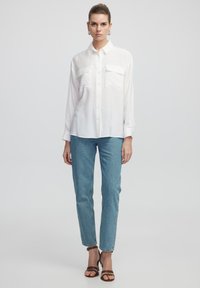 Camisa blanca de botón con dos bolsillos en el pecho, combinada con jeans de denim azul claro y sandalias de tacón marrones. Textura de tela suave.