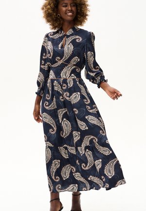 Kobieta w długiej granatowej sukience z beżowymi wzorami paisley, z długimi bufkowatymi rękawami i małym wycięciem typu keyhole, uśmiechająca się i pozująca.