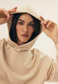 Beżowy hoodie z kapturem na sznurki. Miękka tekstura materiału, luźny krój i widoczne detale szycia. Model trzyma kaptur obiema rękami.