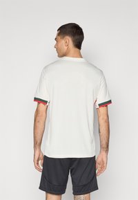 Maglietta sportiva a maniche corte bianca con dettagli verdi e rossi sulle maniche, superficie texturizzata, abbinata a pantaloni corti scuri.
