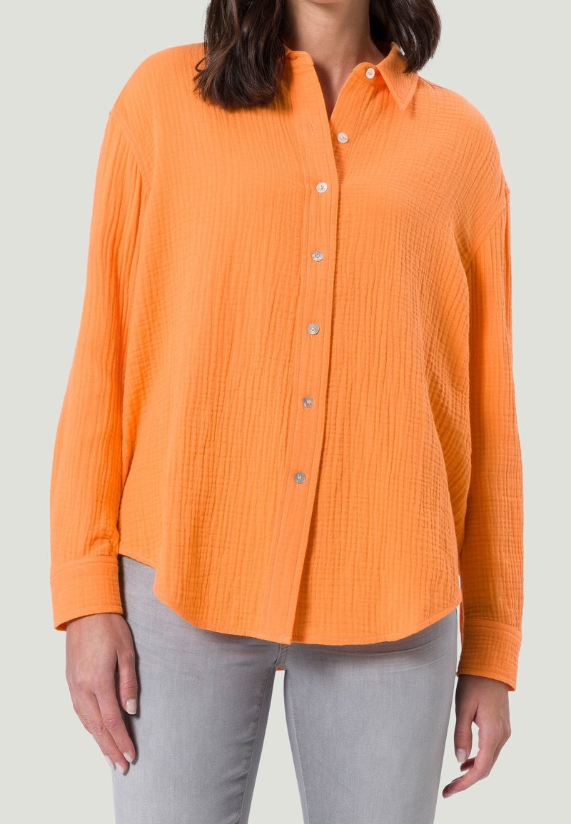 Chemise orange à boutons en tissu texturé, avec un col, des manches longues et des accents de boutons en métal argenté. Assortie avec un jean gris.