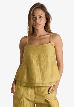 Giovane donna che indossa una canottiera di lino gialla larga e pantaloni abbinati, in piedi con le mani nelle tasche su uno sfondo semplice.