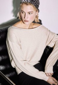 Mlada ženska, oblečena v bež pulover z odkritimi rameni in črno-belo rutko s pikami, sedi na črnem usnjenem stolu.