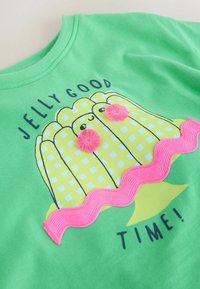 Vihreä paita, jossa keltainen ruudullinen hyytelöjälkiruokagrafiikka, vaaleanpunaiset teksturoidut tupsut ja somisteet sekä teksti "Jelly Good Time!"