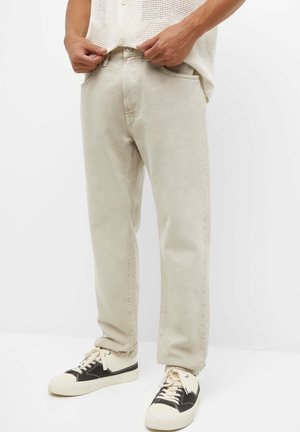 Personne portant un pantalon beige droit et des baskets noir et blanc, ajustant la taille, sur fond blanc uni.
