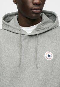 Γκρι φούτερ με κουκούλα από μαλακό ύφασμα, με κυκλικό λογότυπο Converse All Star στο στή θος με κόκκινες και μπλε λεπτομέρειες.