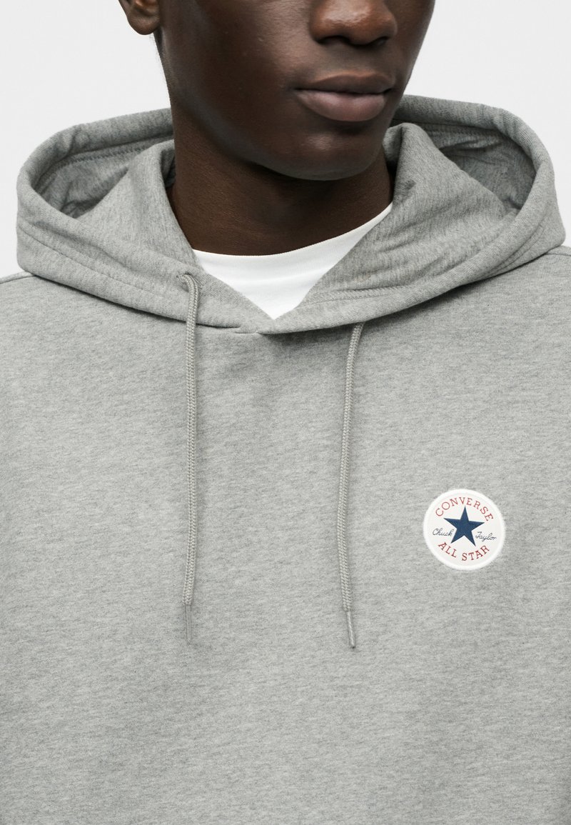 Grå hættetrøje lavet af blødt stof, med en cirkulær Converse All Star-logo på brystet med røde og blå accenter.