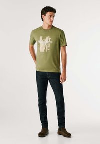 Olijfgroen katoenen T-shirt met een grafische print en de tekst "Pepe Jeans", gecombineerd met donkerblauwe denim jeans en bruine suède enkellaarsjes.