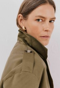Oliwkowy trench z wysokim kołnierzem, z detalami w postaci guzików na ramionach, sparowany z tylko teksturowanymi złotymi kolczykami wkrętami. Gładka tkanina, minimalistyczny design.