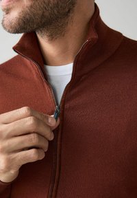 Rozsda színű, cipzáros pulóver kötött anyagból, magas nyakkal és fém cipzár részlettel. Az anyag sima és testhezálló hatású.