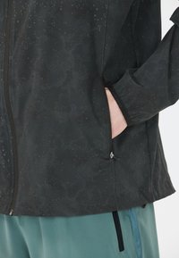 Schwarze wasserdichte Jacke mit strukturiertem Muster und Reißverschlusstasche. Verfügt über elastische Bündchen und ein leichtes, atmungsaktives Material.