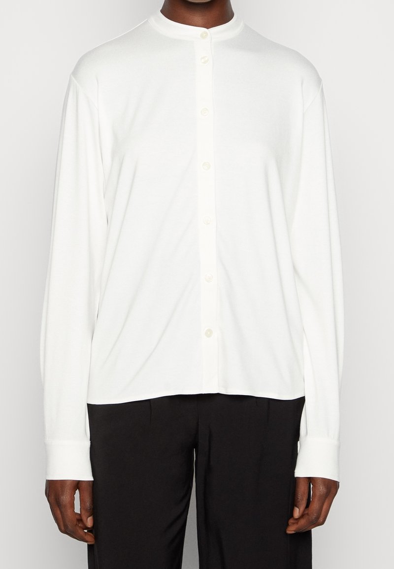 Homme portant une chemise blanche à manches longues boutonnée avec un col rond et un pantalon noir, debout devant un fond uni.