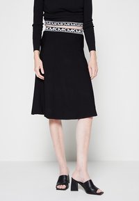 Gonna nera a maglia con vita aderente e orlo svasato, caratterizzata da una fascia a contrasto con il logo "MK". Abbinata a mule nero open-toe con tacco.