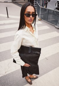 Sac en toile en suede noir avec deux poignées supérieures, agrémenté d'une boucle, porté avec une chemise blanche à boutons et une jupe noire, sur un passage piéton.