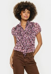 Violetinė, rožinė ir juoda gėlėta bluzelė su raukinėta apykakle, trumpomis rankovėmis ir kaklo raišteliu. Dėvima su rudomis kelnėmis.