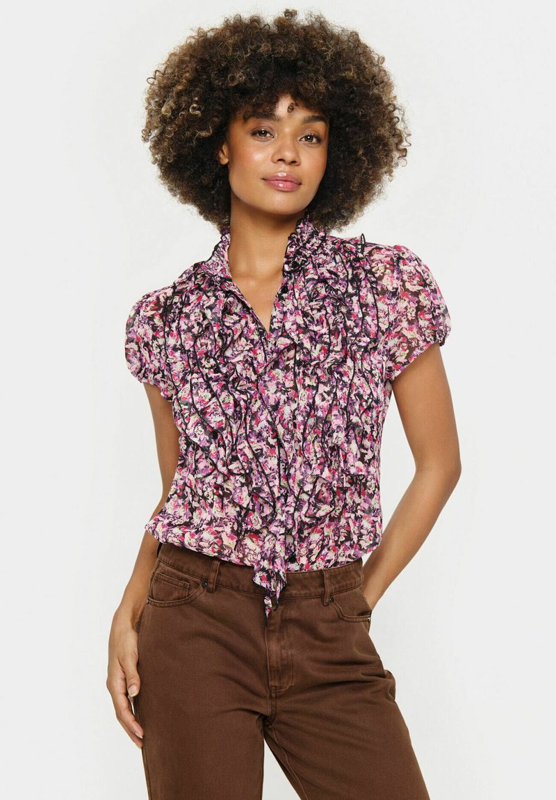 Violetinė, rožinė ir juoda gėlėta bluzelė su raukinėta apykakle, trumpomis rankovėmis ir kaklo raišteliu. Dėvima su rudomis kelnėmis.