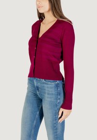 Cardigan aderente con scollo a V profondo in magenta scuro, caratterizzato da polsini a coste e un motivo a righe testurizzato. Abbinato a jeans blu chiaro.