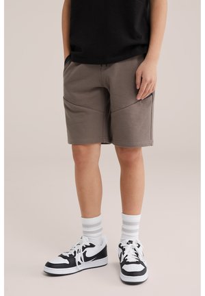 Niño con pantalones cortos grises hasta la rodilla, calcetas blancas con rayas grises y zapatillas Nike en blanco y negro, de pie con las manos en los bolsillos.