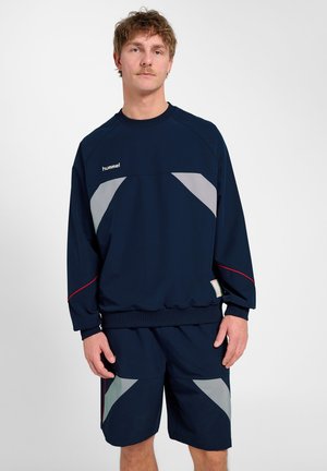 Junger Mann in marineblauer Sportkleidung mit grauen und roten Akzenten, der vor einem schlichten weißen Hintergrund steht.