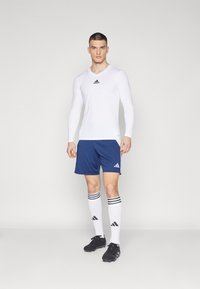 Weißes langärmliges Sportoberteil mit V-Ausschnitt, blaue Shorts mit Seitenstreifen, weiße Socken mit schwarzen Streifen und schwarze Sportschuhe.