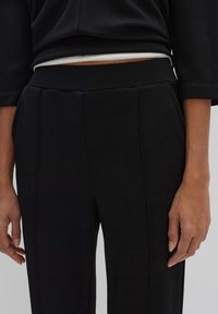 Zwarte gestructureerde broek met een brede tailleband, verticale naden en zijzakken, gecombineerd met een donker topje. Handen zichtbaar aan de zijkanten.