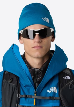 Młoda osoba nosząca niebieską czapkę The North Face, niebieską kurtkę, czarną puchową kamizelkę, czarny plecak i ciemne okulary przeciwsłoneczne w białej oprawie.