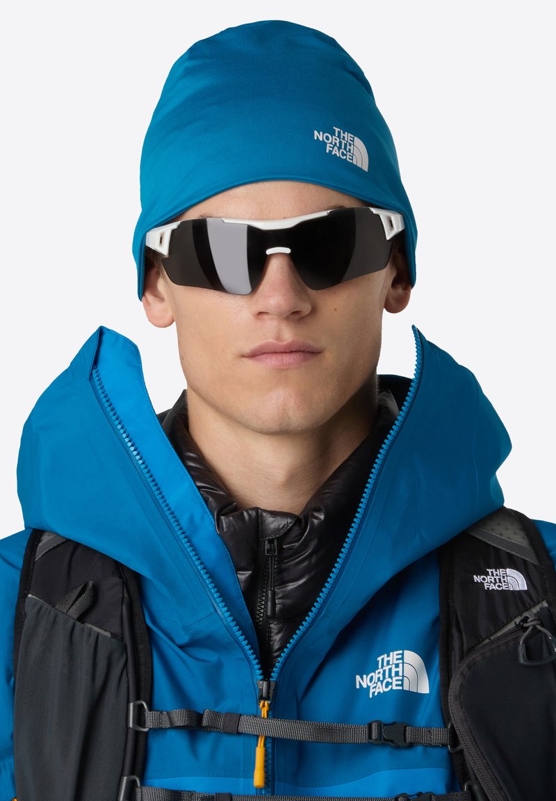 Młoda osoba nosząca niebieską czapkę The North Face, niebieską kurtkę, czarną puchową kamizelkę, czarny plecak i ciemne okulary przeciwsłoneczne w białej oprawie.