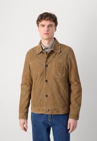 Tan suede jacket with a button front, point collar, and a chest pocket. Features a smooth texture and relaxed fit, paired with blue jeans.