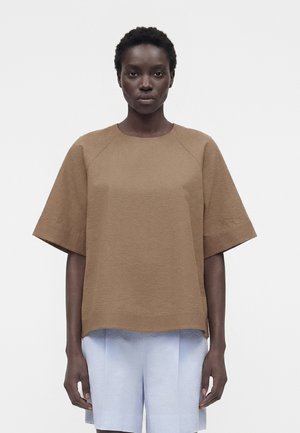 Femme aux cheveux courts et bouclés portant un haut marron ample à manches courtes et un short bleu clair, debout devant un fond blanc uni.