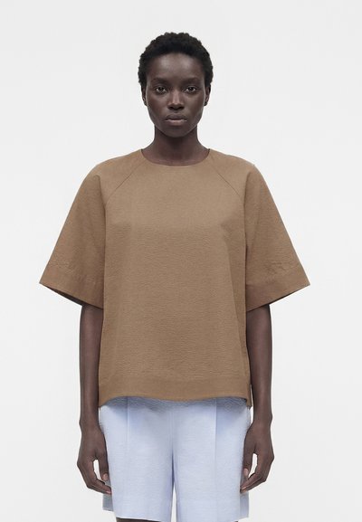 Femme aux cheveux courts et bouclés portant un haut marron ample à manches courtes et un short bleu clair, debout devant un fond blanc uni.