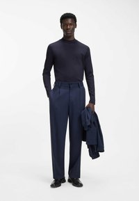 Marineblauer Rollkragenpullover, schlanke Hose und passender Blazer. Glatter Stoff mit dezentem Glanz. Schuhe sind schwarz mit polierter Oberfläche.