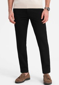 Schwarze, enganliegende Hose mit einer glatten Textur und geradem Schnitt. Getragen mit braunen Wildleder-Lederschuhen. Einfaches Design, ohne sichtbare Muster.