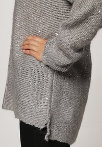 Harmaa neulepusero, jossa on metalliset paljetit, löysä istuvuus, pitkät hihat, teksturoitu kangas ja sivuaukot alahelmassa.