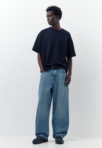 Bărbat purtând un tricou negru supradimensionat, blugi evazați de culoare albastru deschis cu curea neagră și pantofi negri, stând pe un fundal alb simplu.