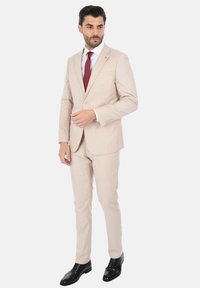 Completo beige con un design aderente, chiusura a un bottone e rever a punta. Abbinato a una camicia bianca e una cravatta rossa, completato da scarpe nere.