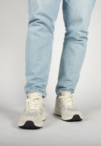 Béžové a šedé tenisky z materiálů síťoviny a semiše, s bílými tkaničkami a černou gumovou podrážkou, nošené s světle modrými džíny.