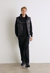 Gilet imbottito nero sopra una felpa scura, abbinato a pantaloni cargo neri e sneakers nere. Il gilet presenta un colletto alto e chiusura con zip.