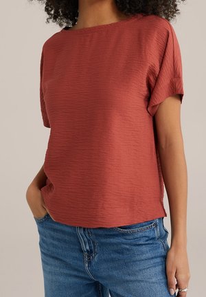 Frau trägt eine strukturierte kurzärmelige Bluse in Rostfarbe, die leicht in blaue Jeans gesteckt ist, mit einer Hand in der Tasche.