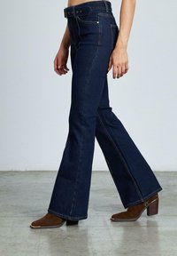 Jeans a zampa d'elefante in denim blu scuro con vita regolabile, cuciture dettagliate e vita media, abbinati a stivaletti marroni con tacco.