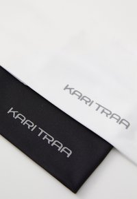 Deux bandeaux en tissu, l'un blanc et l'autre noir, tous deux avec une texture lisse. Chacun présente le nom de la marque "KARI TRAA" en lettres argentées.