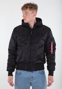 Black bomber jacket with a hood, made of shiny nylon. Features ribbed cuffs, waistband, and a red tag on the sleeve with text.