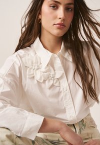 Frau mit langem braunem Haar trägt weißes Hemd mit Knöpfen und zwei großen Stoffblumen auf der Schulter sowie hellfarbene Hosen.