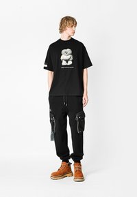 Camiseta negra de gran tamaño con un estampado gráfico, combinada con pantalones cargo negros con bolsillos con cremallera y botas marrones con cordones.