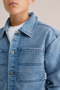 Šviesiai mėlyna džinsinė striukė su apykakle, dviem priekinėmis kišenėmis ir sidabriniais sagčių detalėmis. Yra matoma siuvimo linija ir etikete, pažymėta "BLUE RIDGE."