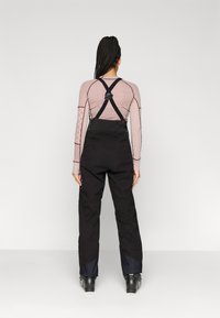 Melnas kombinezonas ar regulējamiem melniem siksnām, aprīkots ar metāla aksesuāriem, kombinēts ar rakstainu rozā un baltā krāsā garām piedurknēm topu.