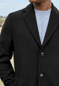 Abrigo de lana negro con cuello de muesca, que presenta tres botones y un suave suéter de punto gris debajo. Textura suave, diseño a medida.