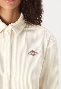 Camisa de pana color crema con cuello, que presenta un logotipo bordado marrón. Superficie texturizada con patrón acanalado.